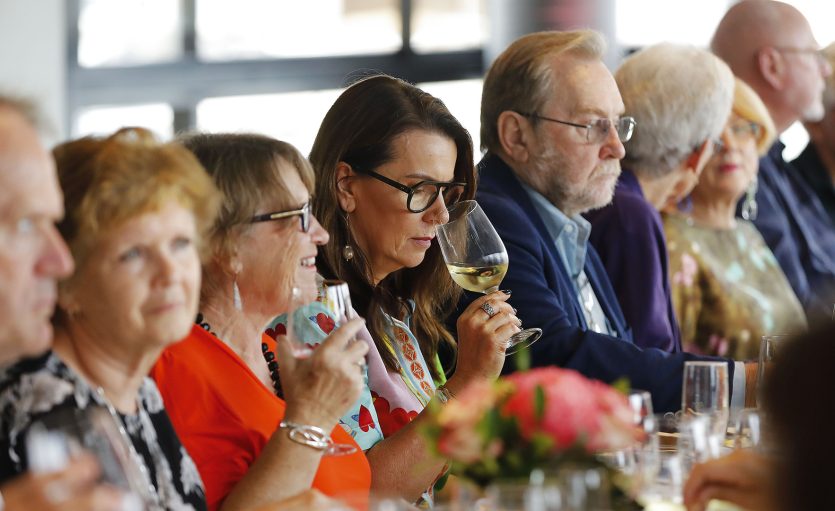A group of people sit together at a long table, enjoying food and drinks. A woman in the centre smells a glass of white wine. The mood appears social and lively, with bright indoor lighting and floral decorations.