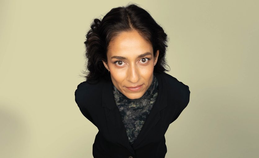 A woman with dark wavy hair and a neutral expression stands against a plain light-coloured background, wearing a dark blazer and patterned top, looking up directly at the camera.