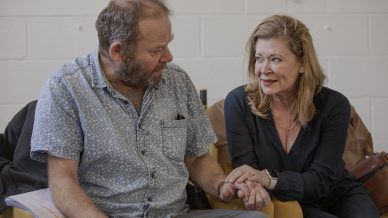 A man and a woman sit closely together indoors, holding hands and having an earnest conversation. The woman looks intently at the man, who is holding some papers in his lap. Both appear thoughtful and engaged.