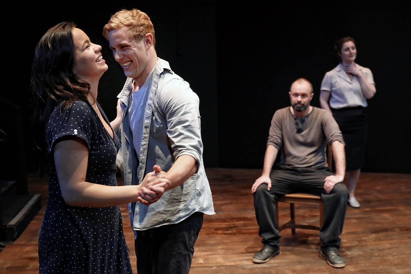 Two people stand and smile at each other whilst holding hands in the foreground. In the background, a man sits on a stool looking serious, and a woman stands nearby, watching the pair. The scene takes place on a wooden stage.