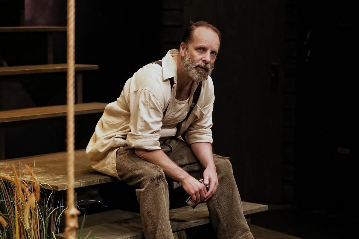 A middle-aged man with a beard, wearing a cream shirt and brown trousers, sits on wooden steps indoors, looking thoughtful with his hands clasped. The setting appears rustic, with plants and a rope nearby.