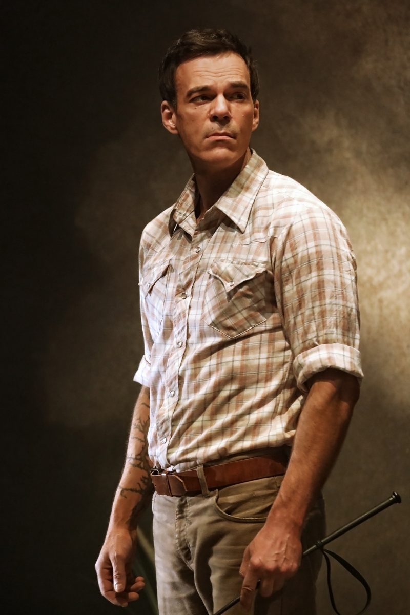 A man in a checked shirt and khaki trousers stands against a dark, textured background, looking off to the side with a serious expression. He holds a riding whip in one hand and has a tattoo on his right forearm.