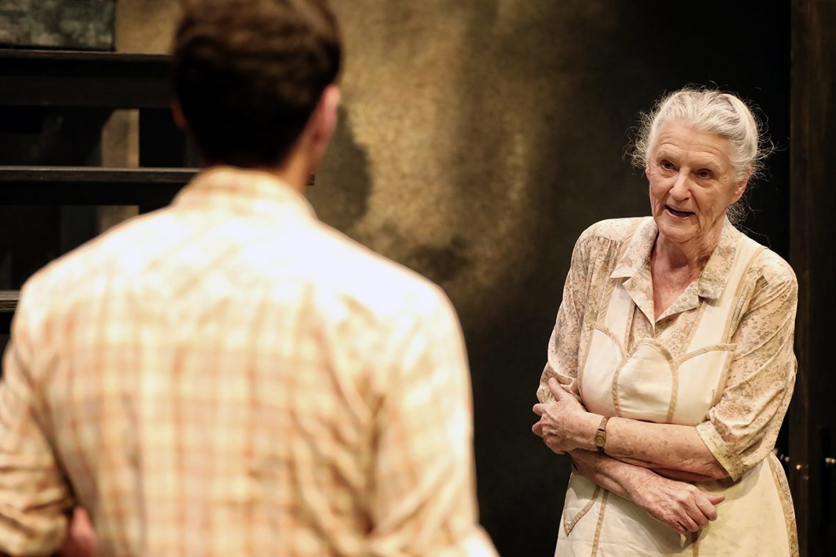 An elderly woman with grey hair in a bun, wearing a light-coloured dress and apron, stands with arms crossed, speaking to a younger man with short hair and a checked shirt, whose back is to the camera.