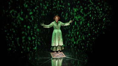 A woman in a green dressing gown stands on a transparent circular platform, arms outstretched. Green, glowing light patterns are projected around her, creating an ethereal atmosphere.