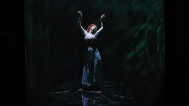 A person stands alone on a dimly lit stage with arms raised, dressed in dramatic clothing and illuminated by a spotlight against a dark, mysterious green-blue backdrop.