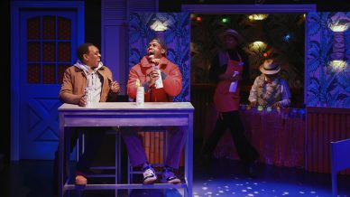 Two men sit laughing at a table with drinks in a colourful, tropical-themed bar. Behind them, a barman in a red apron serves a customer, and island-style decorations create a festive atmosphere under warm lighting.