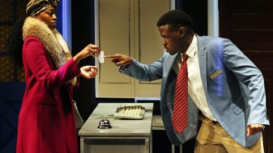 A woman in a red coat and fur collar stands by a desk, handing a card to a man in a blue blazer and striped tie who is reaching out to take it. They are in an office-like setting with cupboards and a bell on the desk.