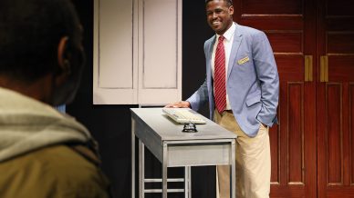 A man in a blue blazer, red striped tie, and khaki trousers stands smiling behind a metal front desk with a bell on it, facing another person whose back is to the camera. Wooden doors and lockers are in the background.
