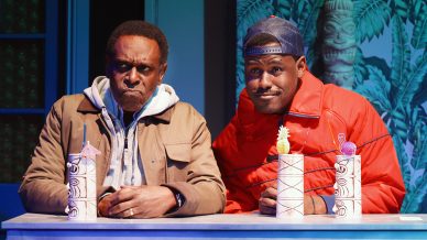 Two men sit at a bar with colourful tiki drinks. The man on the left wears a brown jacket and looks serious, while the man on the right wears a red puffer jacket and cap, smiling slightly. Tropical decor is in the background.