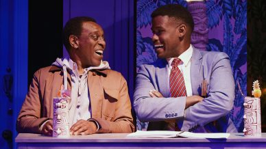 Two men sit at a counter smiling at each other. One wears a brown jacket and hoodie, the other a light blue suit and red striped tie. Tropical-themed drinks are on the counter and a purple-lit background with leafy wallpaper is visible.