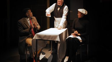 Three people are on stage: a man in a waiter’s uniform stands by a table, gesturing with his fist, while a man in a coat and tie and a woman in a beret and black outfit sit and look at him attentively.