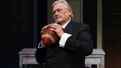 An older man in a dark suit and glasses holds a football close to his chest whilst standing indoors, appearing thoughtful.