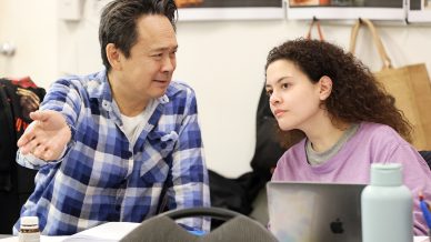 A man in a blue checked shirt gestures while talking to a woman with curly hair in a purple top, who is listening intently near a laptop and water bottle in a classroom or meeting room.