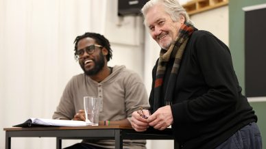 Two men sit together at a table, smiling and laughing. One is seated with a notebook and a glass of water in front of him, while the other leans forward in a scarf. The background shows a classroom or meeting space.