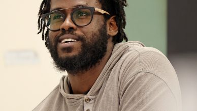 A man with dreadlocks, glasses, and a beard sits at a table with papers, wearing a beige henley shirt and smiling slightly. The background is a classroom setting with a green chalkboard.