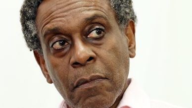 A middle-aged man with short curly grey hair, wearing a light pink button-up shirt, looks to the side with a serious and slightly concerned expression. The background is plain and white.