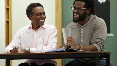 Two men sit at a table, smiling and laughing together. Both have empty glasses in front of them, with one man holding his glass. There is a book or script on the table, and a green chalkboard is in the background.
