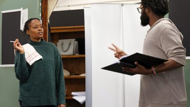 Two people rehearse a scene indoors. One holds a towel and looks thoughtful, while the other gestures with two fingers and reads from a script. Shelves and a partition are in the background.