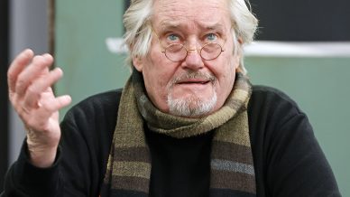 An older man with grey hair, a beard, and glasses sits on a chair. He wears a dark jumper, a striped scarf, and holds a cigarette. He gestures with one hand and looks slightly upward.