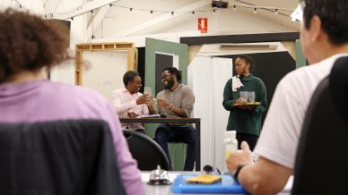 Four people are in a bright room: two men sit at a table talking and laughing, a woman stands holding a tray, and another person sits in the foreground, facing away from the camera.