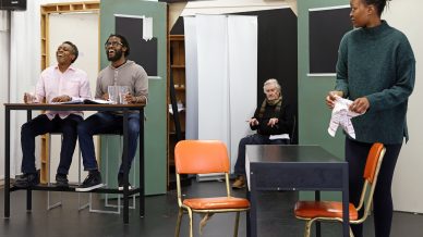 Four people rehearse a scene on a stage set. Two men sit at a high table on the left, laughing. An older man sits in the background, and a woman stands by a table on the right, holding a cloth and looking over.
