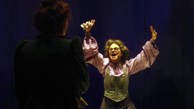A woman wearing glasses, a frilled blouse, and an apron holds her arms up excitedly while facing another person in dim stage lighting against a dark blue background.