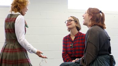 Three women are talking and laughing together indoors. One is standing, holding spectacles and gesturing, while the other two sit, smiling and engaged in conversation against a white brick wall with bright windows.