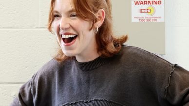 A young woman with red hair smiles widely, standing with her hands on her hips. She wears a dark jumper over a black top and a green skirt, and stands in front of a white wall with a warning sign behind her.