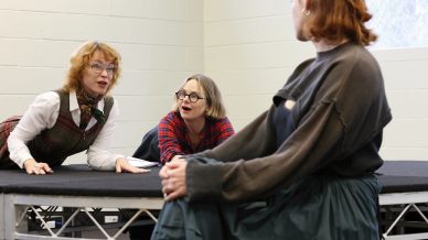 Three women are indoors; two are on a raised platform, one lying down and the other leaning forward, both looking intently at a third woman who sits facing them with her hands clasped in her lap.