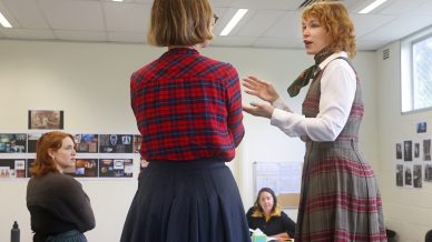 Two women stand and talk in a classroom; one gestures while speaking. Another woman sits nearby, listening, and a fourth person is at the back, working at a desk. Classroom walls have photos and papers displayed.