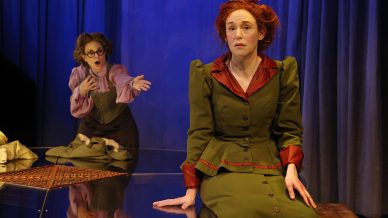 Two women in period costumes perform on stage; one sits on a reflective piano looking pensive, whilst the other stands behind her, gesturing dramatically with outstretched arms. Blue curtains hang in the background.