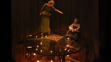 Two actors on a circular, glass stage with warm lighting; one stands and points at the other, who is seated. Both wear vintage-style costumes, surrounded by a dramatic, dimly lit setting.