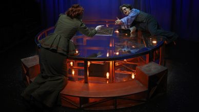 Two people in fancy dress reach towards each other across a large, round, glass table with lights underneath, on a dark stage with blue curtains in the background.