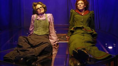 Two people in vintage-style costumes sit side by side on a glass surface, under blue lighting. One wears glasses, a purple blouse, and grey trousers; the other wears a green dress with red trim. Both look upward with relaxed expressions.