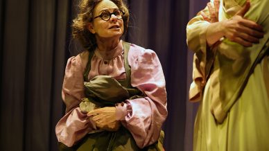 An older woman in glasses and a pink blouse holds a bundle of clothes and looks up at another woman in a pale dress, whose arms are folded. They are on a dimly lit stage with dark curtains in the background.